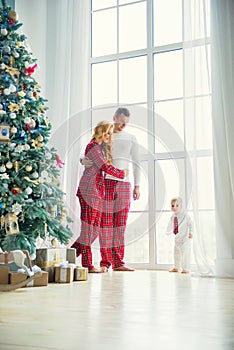 Happy family in plaid pajamas near the big window in the living room with a Christmas tree. Mother, father and little son waiting