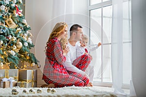 Happy family in plaid pajamas near the window in the living room with a Christmas tree. Mother, father and little son waiting for