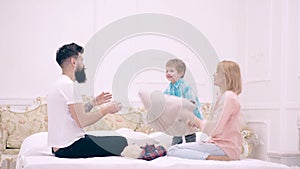 Happy family playing on the bed and throwing pillows. Father, mother and son jump with cushions on the bed.