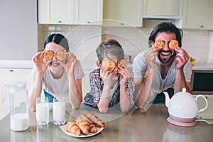 Happy family are playing with rolls. They pyt them on eyes and smiling. Family are leaning to table. There are kettle