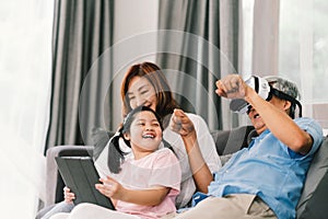 Happy family playing Virtual Reality game together, young kid girl using digital tablet, grandfather wearing VR headset