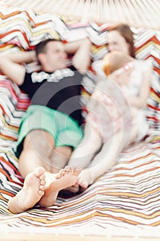 Happy family portrait, young hipster father and mother lying in a hammock with their baby daughter, focus on feet, blurred