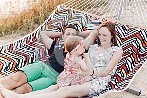 Happy family portrait, young hipster father and mother lying in a hammock with their baby daughter, summer vacation family trip