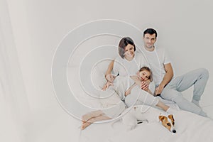 Happy family pose on white bed during weekend. Father, mother, their daughter and dog feel comfort of being in bedroom, embrace