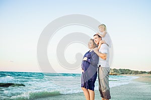Happy family- pregnant mother, father and daughter embracing each other and watching ahead to the sea during walk on the beach. Re