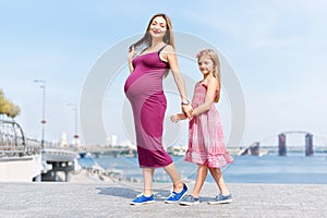 Happy family, pregnant mother and her daughter little girl child walking and hugging on the embankment in the summer day