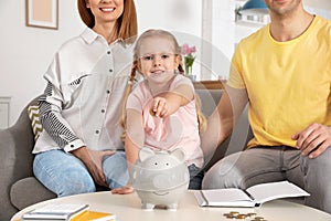 Happy family putting coin into piggy bank at table. Saving money