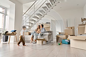 Happy family relaxing on couch on moving day with kid son playing in new home .