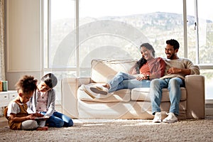 Happy family relaxing at home. Smiling young parents with girls on a sofa, watching their cute little children play and