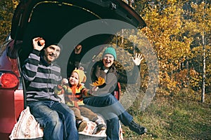 Happy family resting after day spending outdoor in autumn park. Father, mother and child sitting inside car trunk, smiling and