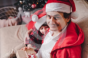 Happy family in Santa hat, father and child son giving Christmas gift at home. Sitting on a couch in the living room