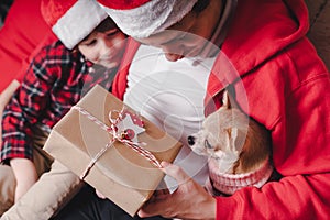 Happy family in Santa hat, father and child son giving Christmas gift at home. Sitting on a couch in the living room with puppy