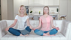 Happy family senior mother and grownup daughter on the sofa practicing yoga meditation at home. Adult woman and her