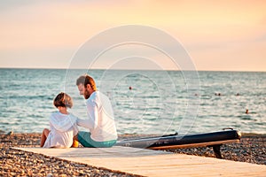 Happy family sits at the pebble beach and relax after swimming. Back view. Sunset light. Copy space. Summer vacation at