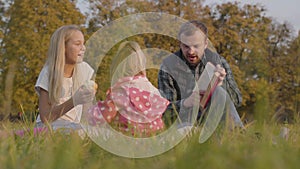 Happy family sitting on the autumn meadow, eating apples and reading the book in red cover. Bearded caucasian man having