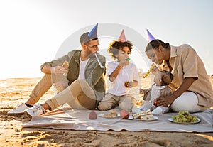 Happy family sitting on blanket at the beach, wearing party hats and blowing whistles, celebrating birthday outdoors