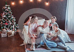 Happy family sitting on the floor by the bed in the new year interior. Dad is reading a book. Children laugh. Cozy room