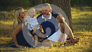 Happy family is sitting on the grass and doing selfie with a baby at sunset in the park. Father and mother take pictures