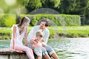Happy family sitting on jetty on lake or pond