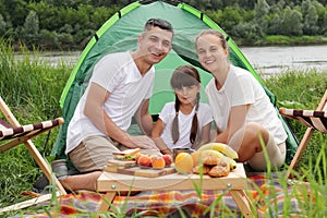 Happy family sitting near tent on the ground having picnic together enjoying tasty fruit and sandwiches, spending happy weekend in
