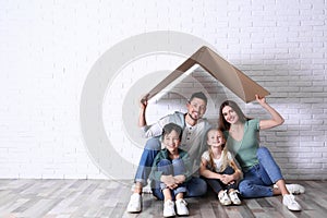 Happy family sitting under cardboard roof near wall, space for text. Insurance concept