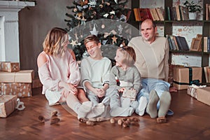 Happy family sitting on the wooden floor in the room next to the Christmas tree. They laugh. On the floor are cones. Warm colour