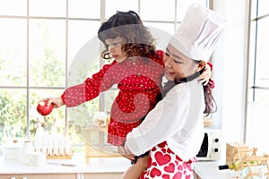 Happy family, smiling beautiful Asian mother wears cute red heart apron and chef hat holds daughter while little girl holds apple