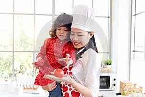 Happy family, smiling beautiful Asian mother wears cute red heart apron and chef hat holds daughter while little girl holds apple