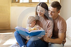 Happy Family Of Three With Cute Infant Son Reading Book Together At Home
