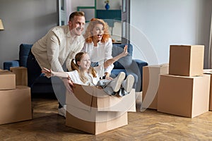 Happy family of three having fun and celebrating moving day, father and mother riding smiling daughter in cardboard box