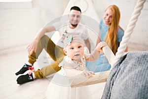 A happy family of three. Mom, dad, child one year old girl in dress play, laugh, smile in bright room. Sunny