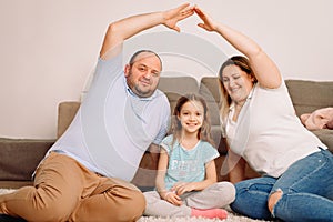 Happy family of three persons sitting together on the carpet with daughter between mother and father making sign of roof