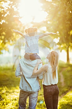 Happy family of three persons walking the grass in the park on sunset. happy daughter flying on father back