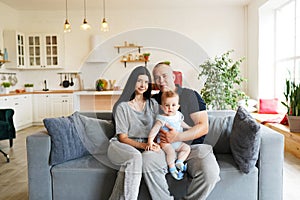 Happy family of three portrait, young parents mother and father and little boy in arms look at camera posing together