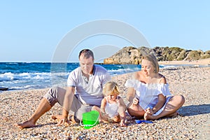 Happy family of three - pregnant mother, father and daughter having fun, playing with sand and shells on the beach. Family vacatio