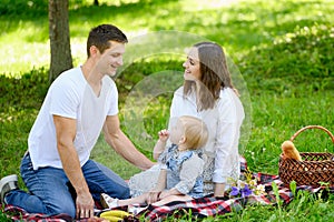 Happy family with toddler having picnic in park in summer. Mom and dad are sitting with child on grass in park, eating