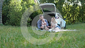 Happy family travels by family car. Picnic outside the city. Father, mother and daughter are sitting on a rug in the