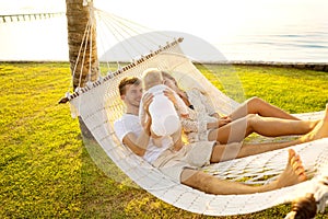 Happy family on a tropical island at sunset lie in a hammock and play with their son