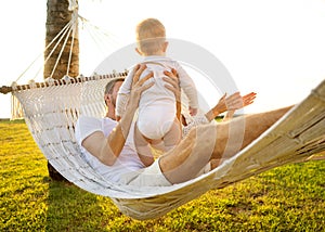 Happy family on a tropical island at sunset lie in a hammock and play with their son