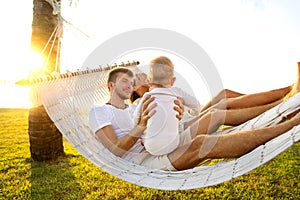 Happy family on a tropical island at sunset lie in a hammock and play with their son