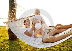 Happy family on a tropical island at sunset lie in a hammock and play with their son