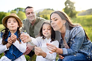 Happy family with two small daughters sitting outdoors in spring nature, blowing dandelion seeds.