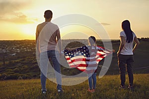 Happy family with usa flag on nature in the evening at sunset. Back view. Mother father and daughter celebrate fun