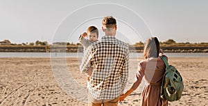 Happy family walking on sandy beach of river. Father, mother holding baby son on hands and going together. Rear view. Family Ties