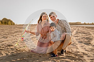 Happy family walking on sandy beach of river. Father, mother holding baby son on hands and playing with kite