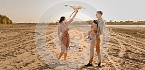 Happy family walking on sandy beach of river. Father, mother holding baby son on hands and playing with kite