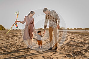 Happy family walking on sandy beach of river. Father, mother holding baby son on hands and playing with kite