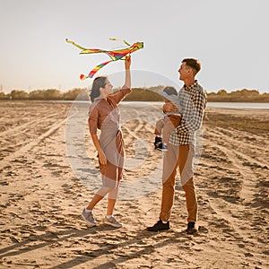 Happy family walking on sandy beach of river. Father, mother holding baby son on hands and playing with kite