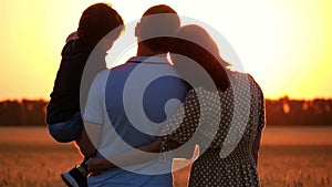 Happy family watching the sunset, standing in a wheat field. A man holding a child in his arms. A woman hugs a man