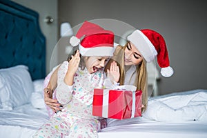 Happy family wearing Santa Claus hats sitting on bed. Mother give and surprised child Santa gift and having fun in Christmas time.